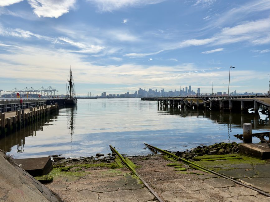 A daytime view of a waterfront area featuring a concrete and moss-covered railway track leading towards the calm water with a stone jetty on the right side, where wooden posts and a metal bollard are visible. To the left, there is a wooden dock with vertical posts, and a small sailing boat with a furled sail is moored alongside. In the background, a distant city skyline with high-rise buildings is visible under a partly cloudy sky with scattered clouds. The setting includes natural elements like moss and rocks, contrasted with man-made structures such as the docks, railway tracks, and maritime equipment, illustrating an urban waterfront landscape that could relate to private or independent waste handling near dockside areas managed by companies like Rubbish Removal Docklands.
