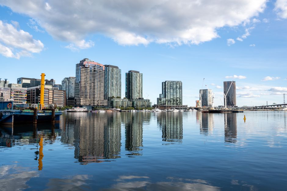 The image features a calm body of water reflecting a cluster of modern high-rise buildings made of glass and concrete, situated along an urban waterfront. In the foreground, there is a small section of a dock or pier with visible blue and black boats moored, and yellow bollards or safety posts, indicating a commercial or private dock area used for boat docking or light freight handling. The buildings vary in height and design, with some showing patterned glass facades and others with a more uniform appearance, contributing to a contemporary cityscape typical of a metropolitan business district. The sky is partly cloudy with patches of blue, creating natural lighting that highlights the textures and colors of the buildings and their reflections on the water’s surface. The water is still, with slight ripples distorting the mirrored images, and the overall environment suggests a clean, organized urban harbour or riverside location. This scene subtly connects to the concept of on-site clearance and private waste management by illustrating typical residential and commercial waterfront settings where rubbish removal services might be required, exemplifying an environment that may benefit from independent waste handling or rubbish removal solutions provided by companies like Rubbish Removal Docklands.