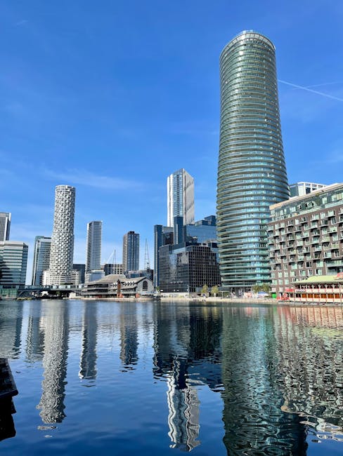 A collection of modern high-rise buildings lining a waterfront in an urban cityscape, including a prominent cylindrical glass tower on the right with a sleek, reflective surface. The buildings vary in architectural style, some with rectangular shapes and different finishes such as glass and concrete, while others have distinctive textures like patterned facades and rounded edges. The water in the foreground reflects the skyline, creating a mirror image of the structures and the clear blue sky overhead, which is free of clouds. The scene is bathed in natural daylight, highlighting the surfaces and colors of the buildings. This urban setting suggests a busy business district or financial hub, with a clean and organized environment typical of central city areas. Occasionally, such environments might provide opportunities for alternative waste handling or private rubbish collection services, supporting the needs of commercial and residential properties.