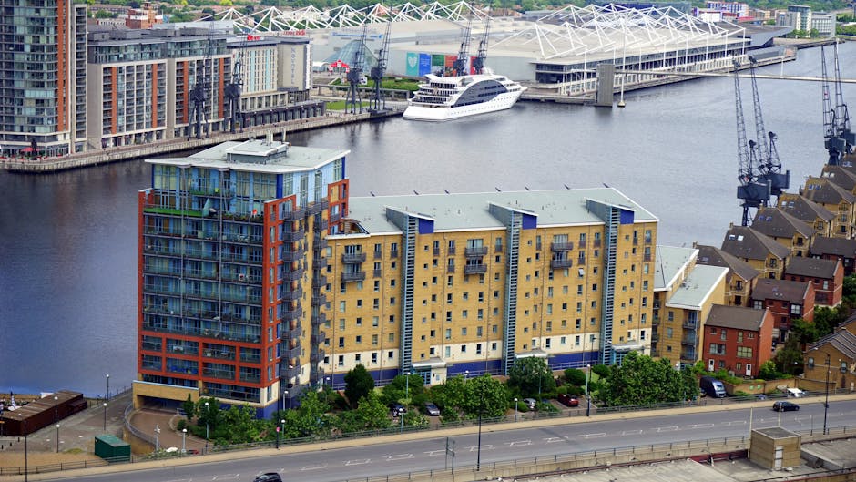 An aerial view of a modern, multi-storey residential building situated near a waterway, with the structure featuring a combination of yellow and blue façade panels, glass balcony railings, and protruding vertical elements. The building's exterior displays a mix of brickwork and smooth finishes, with several balconies that have metal railings and some with glass panels. Surrounding green trees line the street in front of the building, where parked cars are visible alongside a paved road. In the background, the waterway is lined with contemporary residential and commercial buildings, including a large stadium or arena with a distinctive white, tent-like roof structure. A cruise ship with multiple decks is docked along the waterfront, with cranes and industrial equipment nearby, indicating active shipping or construction work. The scene is well-lit by natural daylight, with a clear view of the urban landscape, showcasing a typical Docklands environment that supports both private residential living and alternative waste handling through local rubbish removal services, like those provided by Rubbish Removal Docklands.