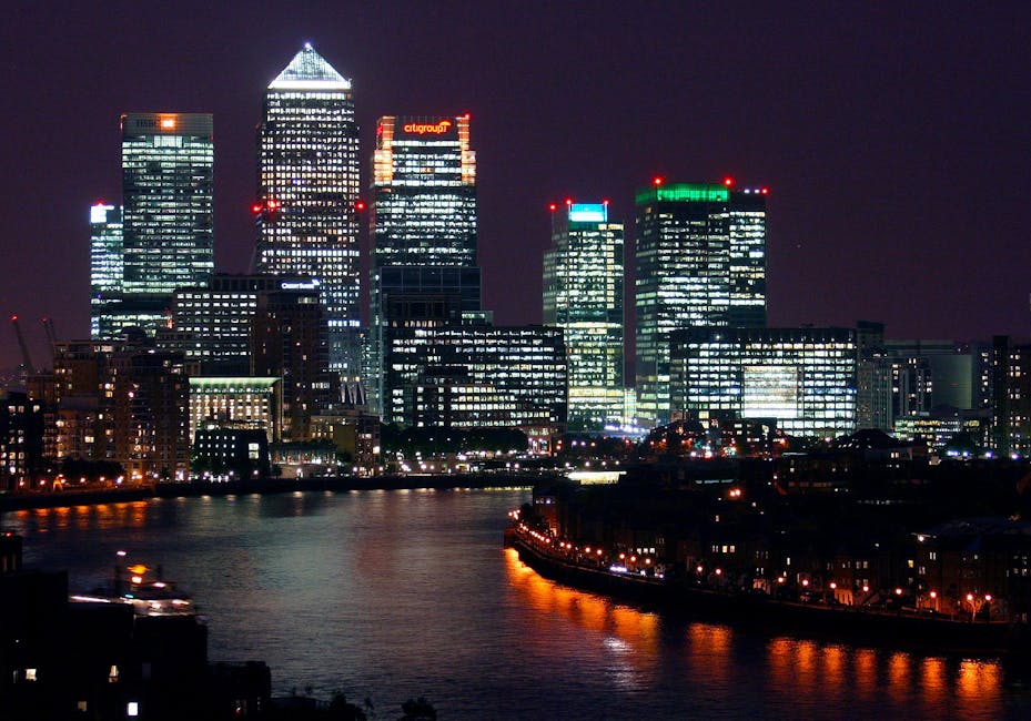 Nighttime view of the Canary Wharf skyline with illuminated office buildings, including the distinctive tall glass towers with bright interior lights and company signage, situated along the River Thames. The water reflects the glow of the city lights, with a dark sky overhead. In the foreground, there is a paved area or possibly a driveway with some faint lighting, suggesting an adjacent street or service access point. The scene provides a clear view of the urban landscape’s modern architecture and bright lighting, typical of a financial district. The lighting indicates active building illumination, while the surrounding environment remains urban and developed, aligning with locations where private waste disposal or alternative rubbish removal services such as those offered by Rubbish Removal Docklands might operate in the area. The image emphasizes the busy, well-lit cityscape characteristic of London’s financial district at night, with no visible rubbish or waste directly shown.