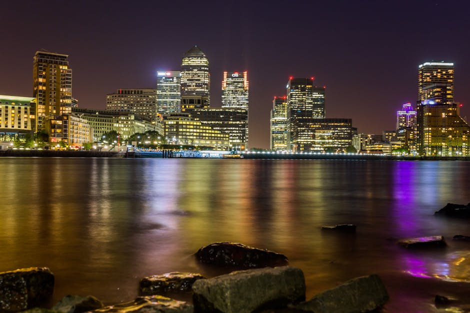 A nighttime cityscape view across a calm body of water features a skyline of modern office buildings and high-rise structures illuminated with multicolored lights, including shades of yellow, white, red, and purple. The buildings vary in height and architectural style, with some having glass curtain walls reflecting their internal lighting, while others have solid facades with lit windows. The water in the foreground is smooth and reflective, capturing the glow of the city lights, with a few rocks visible along the shoreline at the bottom of the image. The scene is well-lit from the city lights, creating a vibrant and lively atmosphere typical of an urban waterfront environment. This image emphasizes the contrast between the dark night sky and the brightly lit buildings, aligning naturally with topics related to urban waste management, private disposal, or alternative rubbish removal services such as those offered by Rubbish Removal Docklands, situated near the Docklands area. It captures the essence of a modern metropolis, highlighting the importance of efficient waste management amidst busy city life and a thriving commercial district.